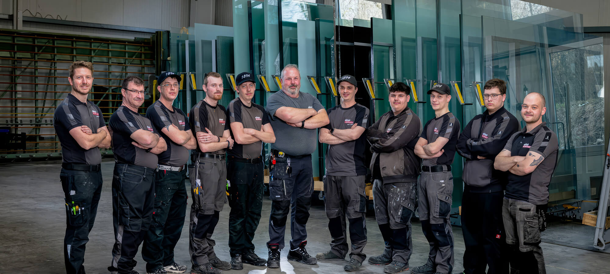 Elf Mitarbeitende eines Glasbetriebs stehen in einer Werkhalle vor großen stehenden Glasscheiben und blicken in die Kamera.